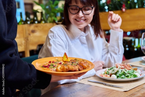 Woman sitting in restaurant  rejoices at plate of cooked food in hands of waiter