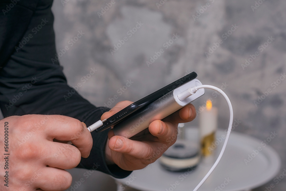 Man holds a power bank in his hands and charges a smartphone against a concrete wall.