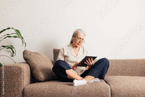 Schilderij op canvas Senior woman sitting on a sofa with crossed legs holding digital tablet