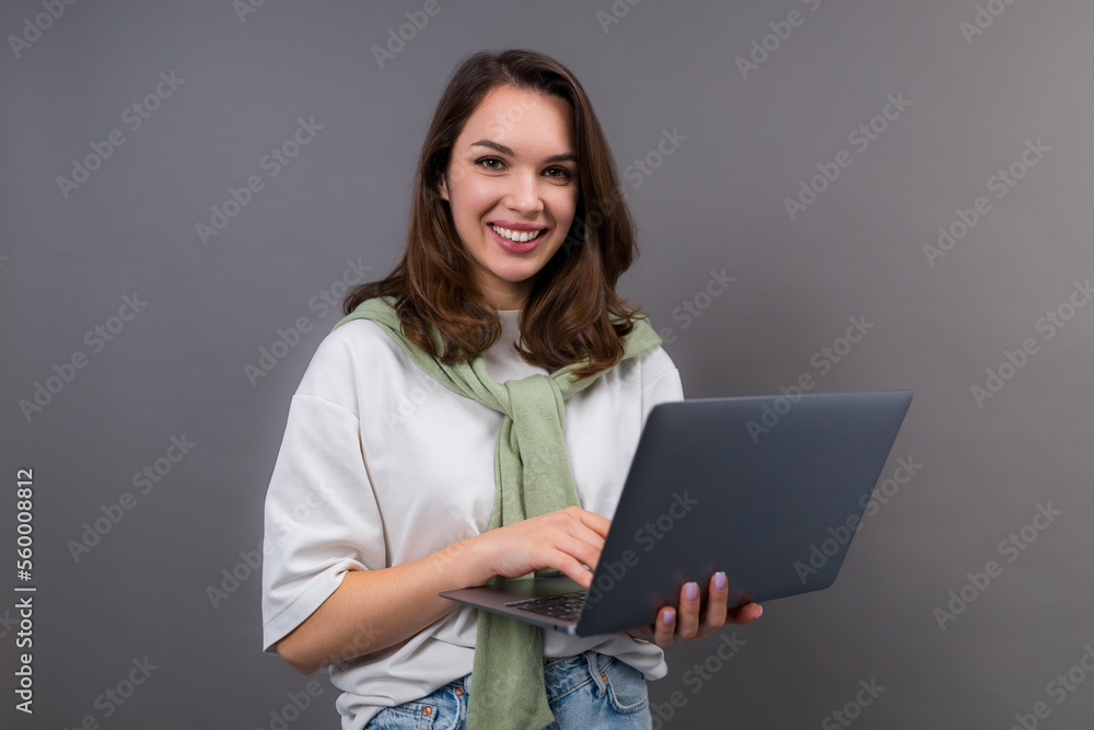 A young smiling woman in a white T-shirt and sweater stands with a laptop on a gray background and looks at the camera. Portrait of a freelancer A young smiling woman in a white T-shirt and sweater stands with a laptop on a gray background and looks at the camera. Portrait of a freelancer