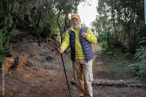 Old smiling senior man enjoying freedom in outdoors walking in a mountain footpath. Elderly active man traveling in a park holding backpack and stick