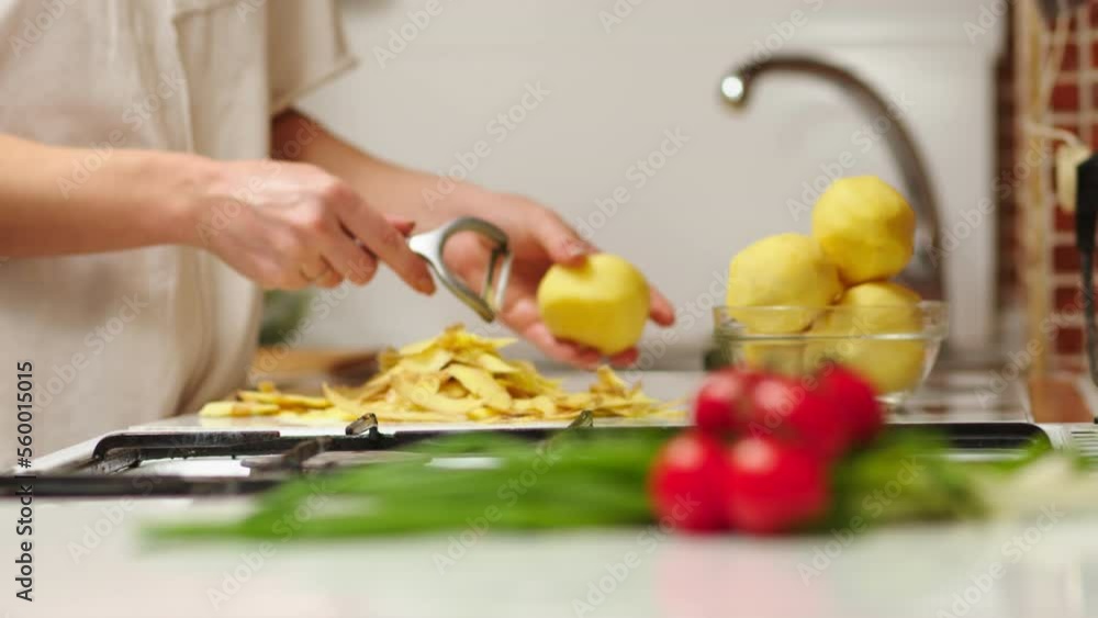 Close up view of woman in white shirt peeling a potato. Peel falling onto cutting board. Lady preparing potatoes to be cut and added to stock pot for dinner. Close up view of woman in white shirt peeling a potato. Peel falling onto cutting board. Lady preparing potatoes to be cut and added to stock pot for dinner.