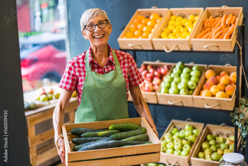 Mature woman works in fruits and vegetables shop. She is holding basket with cucumber.