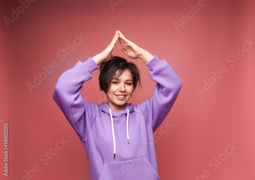 A happy young girl with short black hair makes a gesture of the roof of the house with her hands over her head. The concept of mortgage insurance. isolated on a pink background © inna717