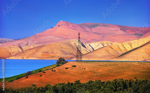 Power line support at the background of barren mountain range, desert, field, bush, evening sky and bright blue water of Al Wahda Dam Lake. Beautiful scenic view in Taounate Province, Morocco, Africa