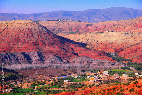 Beautiful desert view, barren multicolored hills in red, pink, purple and yellow colors, little village, few green fields and trees, distant mountains and blue sky in Morocco, North West Africa.
