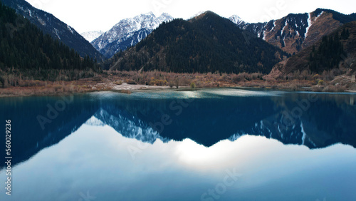 Issyk mountain lake with mirror water at sunset. The color of the water changes before our eyes. There are trees in clear water. Snowy mountains and green hills are visible. Clouds are reflected