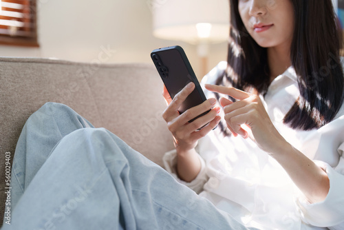 Smiling pretty asian young woman wearing casual clothes using cell phone sitting on sofa at home