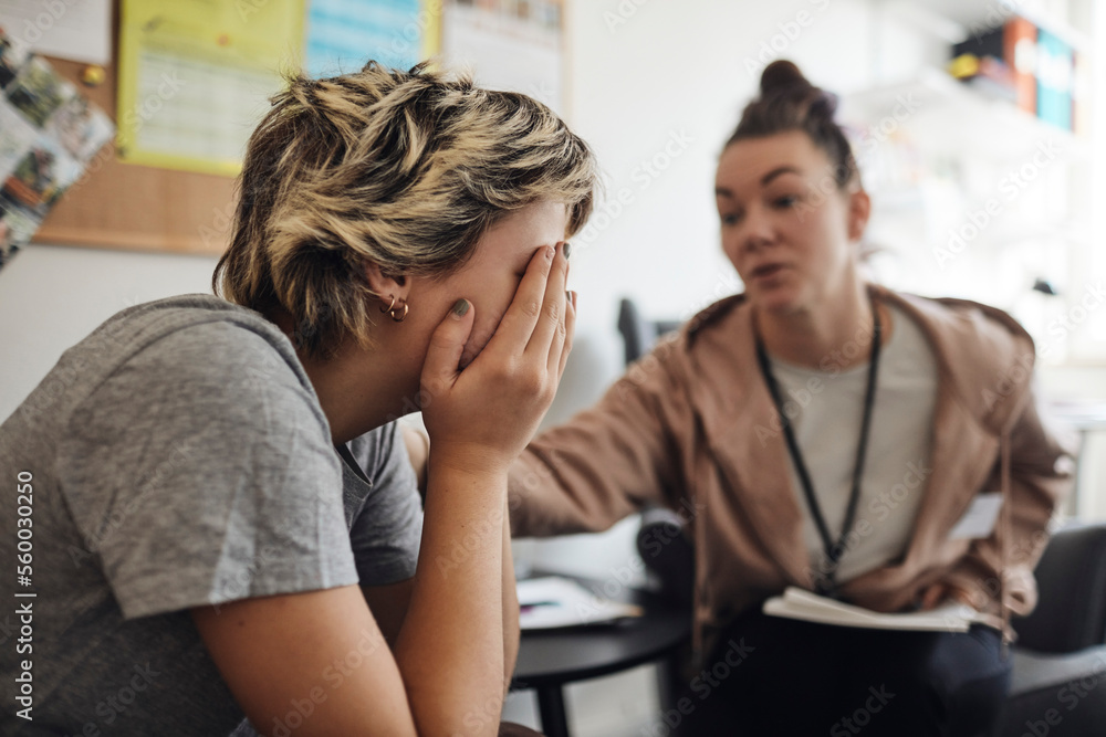 Non-binary counselor consoling female student covering face with hand in school office Non-binary counselor consoling female student covering face with hand in school office