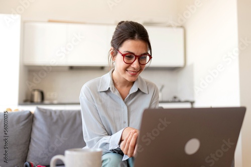 Smiling young woman with laptop communicating with client via video call working at home