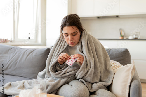 Ill young woman takes pills sitting at home on the sofa under a warm blanket