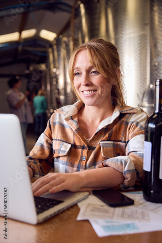 Woman With Laptop Checking Label On Bottle Inside Winery With Storage Tanks