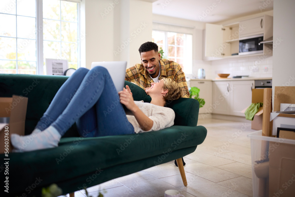 Couple Unpacking Boxes After Moving Into New Home Together As Woman Lies On Sofa Using Laptop Couple Unpacking Boxes After Moving Into New Home Together As Woman Lies On Sofa Using Laptop