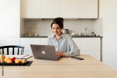 Pretty woman studying online sitting at her desk with her laptop, chatting via video link in a bright apartment