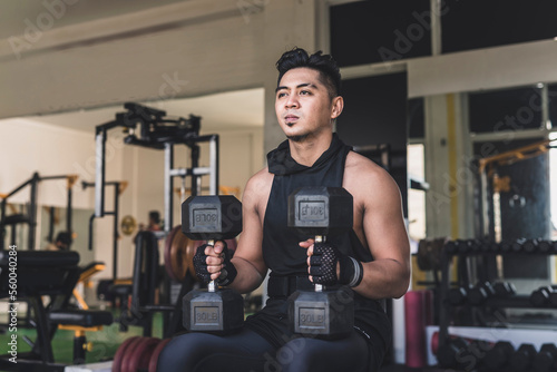 An asian man mentally preparing himself while sitting on an incline bench holding two dumbbells placed on his thighs near the knees preparing to do a set of seated dumbbell shoulder presses.