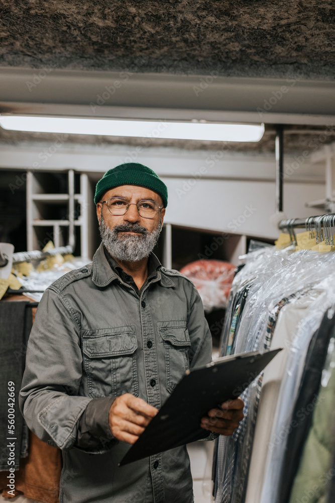 Portrait of bearded tailor with clipboard standing by clothing rack in workshop Portrait of bearded tailor with clipboard standing by clothing rack in workshop