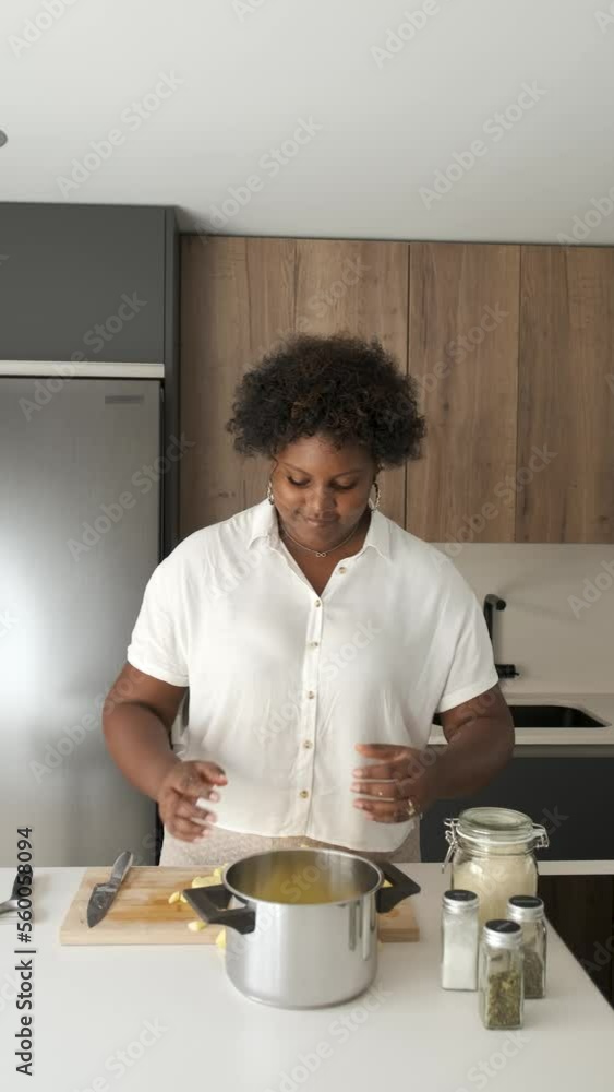 Smiley young cuban woman putting chopped raw potato into a pot to cook it. Smiley young cuban woman putting chopped raw potato into a pot to cook it.