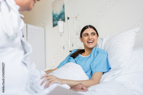 Cheerful patient in gown lying on bed near blurred doctor in clinic.