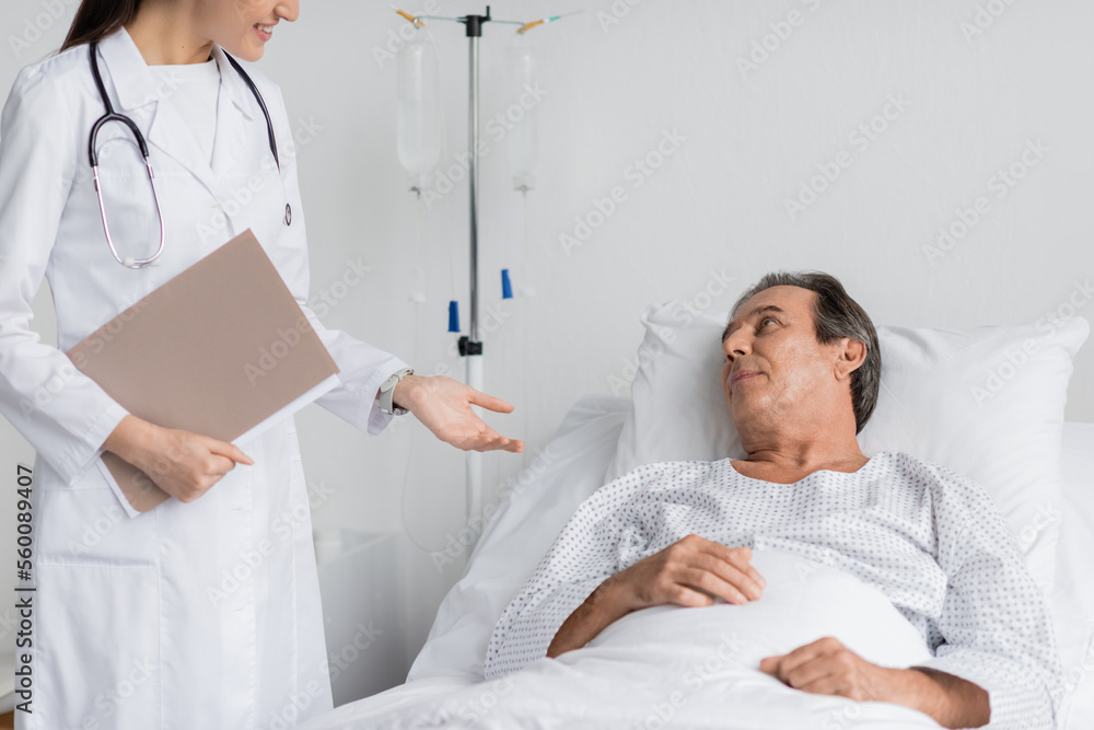 Smiling doctor with paper folder talking to senior patient on bed in clinic.