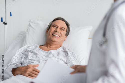 Cheerful patient in gown looking at blurred doctor with paper folder in hospital ward.