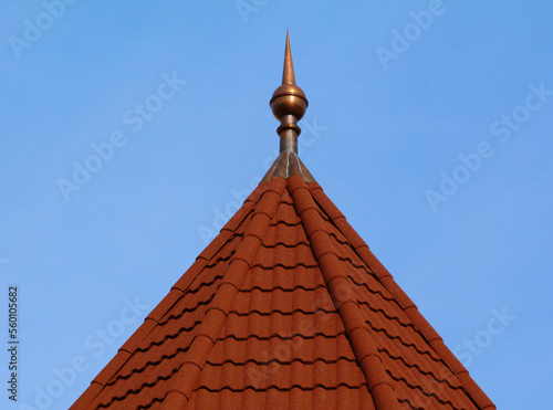 pointy spire on turret with hexagonal sloped building roof top. red clay tile roof. copper spike or spire at the tip. ridge tiles. blue sky background. building materials and construction concept.