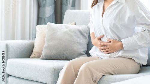 Young woman wearing casual clothes suffering from menstrual pain, feeling sick to her stomach, holding belly, having abdominal cramps during period and lying down on sofa in the living room at home.