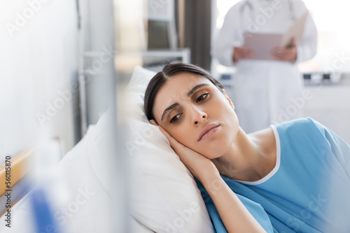 Dissatisfied patient lying on bed near blurred doctor in hospital ward.