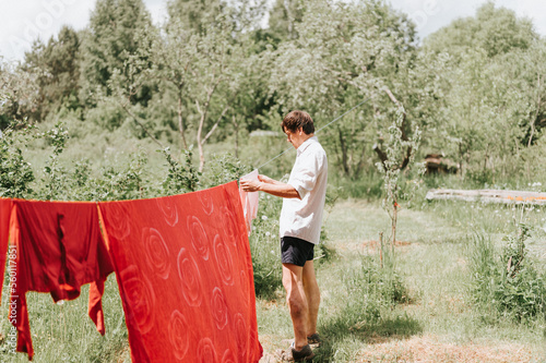 candid young mature choring man chore homework and hanging the laundry to dry on a clothesline on the street in courtyard of village household cottage house. summer, freshness and laundry day photo