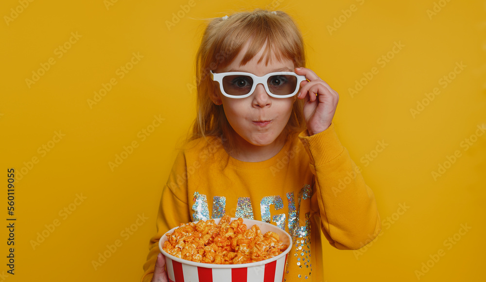 Excited young toddler school girl in 3D glasses eating popcorn, watching interesting tv serial, sport game, film, online social media movie content. Teen female child kid on studio yellow background Excited young toddler school girl in 3D glasses eating popcorn, watching interesting tv serial, sport game, film, online social media movie content. Teen female child kid on studio yellow background