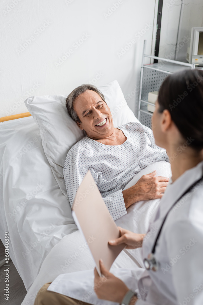 Fototapeta premium Positive senior patient looking at blurred doctor with paper folder in hospital ward.