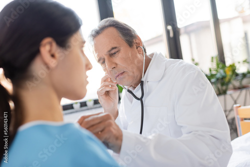 Elderly doctor examining lungs of blurred patient in hospital ward.