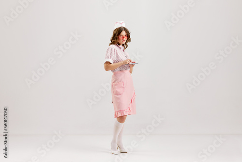 Fototapeta Naklejka Na Ścianę i Meble -  Emotional young woman, retro waitress in american fashion style of 70s, 80s uniform posing over light studio background. Comic photography style