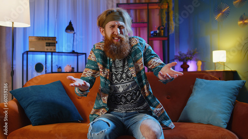 What. Why. Bearded hippie man raising hands in indignant expression asking reason of failure demonstrating disbelief irritation by troubles at home apartment. Young guy in evening room sitting on sofa