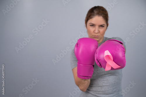 Caucasian woman in pink boxing gloves with a pink ribbon on her chest on a gray background. Fight against breast cancer. 