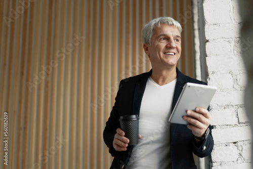 Obraz na plátně A mature gray-haired man at the window takes notes online on a tablet and looks away in a modern office, in casual clothes