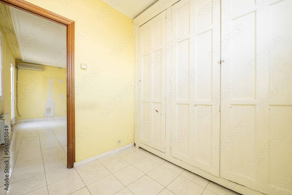 An empty room with walls covered by a large built-in wardrobe with vintage-style white wooden doors and yellow painted walls, a white stoneware floor and a door to another room An empty room with walls covered by a large built-in wardrobe with vintage-style white wooden doors and yellow painted walls, a white stoneware floor and a door to another room