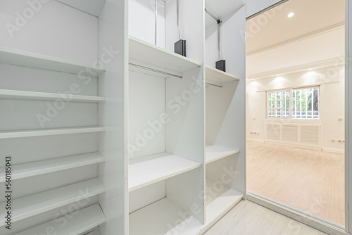 An empty room with white painted walls with access to a dressing room with multiple white marera shelves and French style aluminum windows