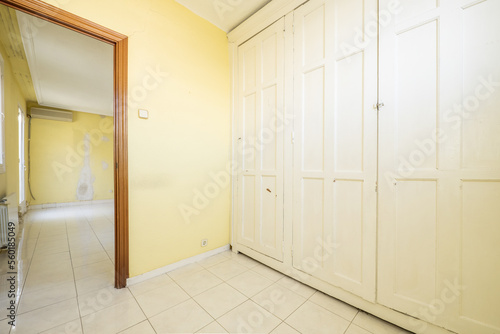 An empty room with walls covered by a large built-in wardrobe with vintage-style white wooden doors and yellow painted walls, a white stoneware floor and a door to another room
