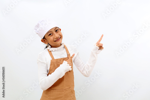 Happy little girl in chef uniform pointing something at blank space. Isolated on white background