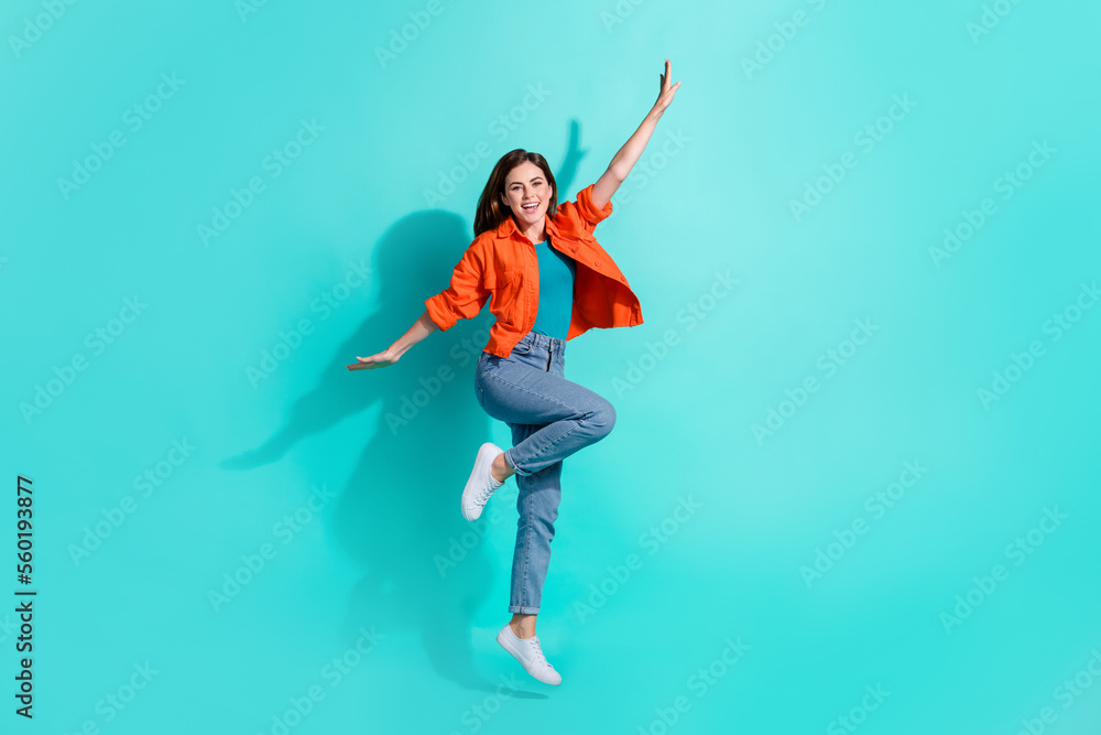 Full length photo of excited pretty girl dressed orange shirt arms sides jumping high isolated turquoise color background