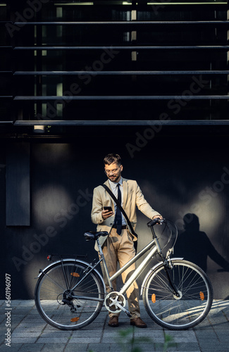 Portrait of business man with bicycle standing in front of office building using smartphone.