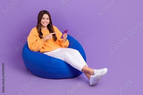 Full length photo of sweet positive girl dressed orange sweatshirt bean bag pointing device empty space isolated purple color background