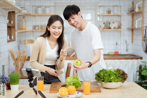 Happy cheerful Asian man and woman talking about cooking and food nutrition in a beautiful decorated kitchen on social network together. Lovely couple making an online live steaming on social media.