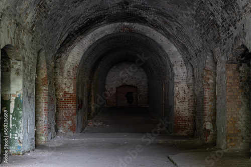 A long dark dimly tunnel of an abandoned military bunker or bomb shelter with broken red brick walls. The arched ceiling is illuminated by daylight from the outside. The floor is dirty and dusty