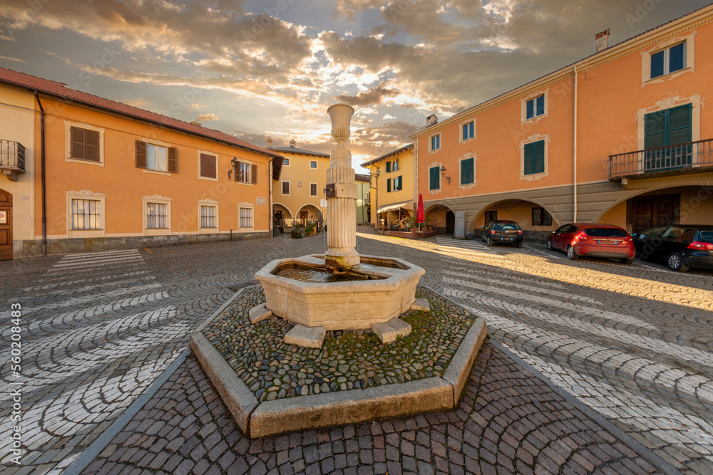 Obraz premium Peveragno, Cuneo, Italy - January 09; 2023: marble fountain with drinking trough on cobblestones in Piazza Santa Maria with ancient palaces decorati with arcade under cloudy sky at sunset