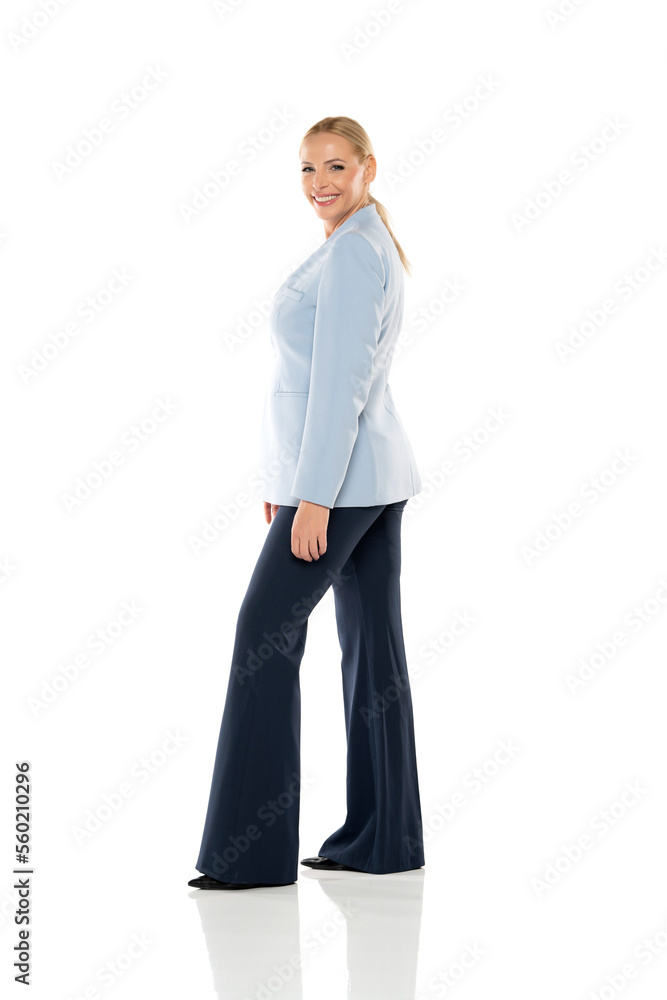 Middle aged senior smiling woman wearing blue jacket and black trousers on a white background. Side view.