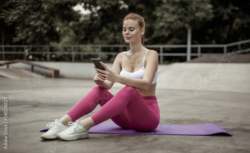 Red-haired fit slim woman using smartphone while sitting on yoga mat outdoors