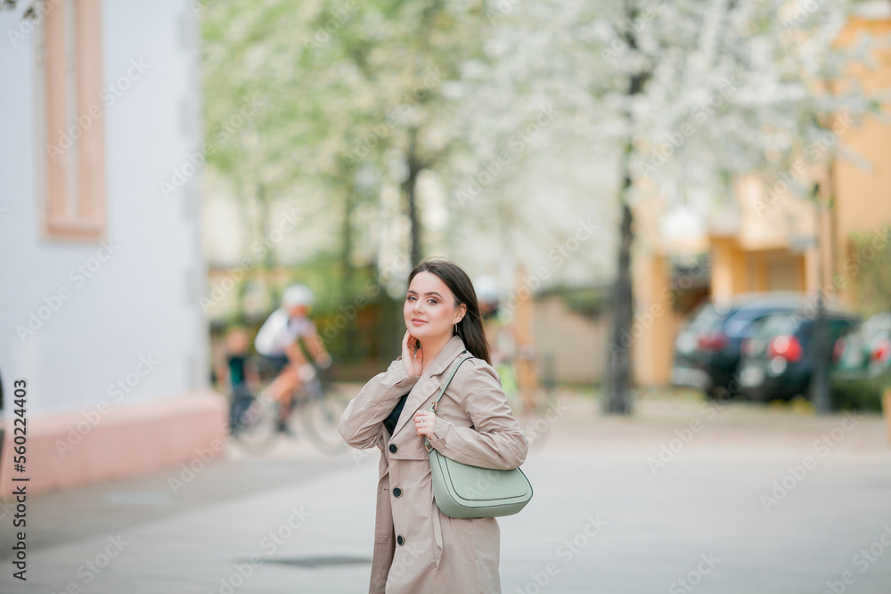 Young beautiful woman 19 years old in stylish clothes. Model with dark hair in casual clothes walks through the spring blooming city. Warm.