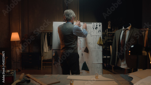 Male mature tailor in luxury designer atelier or tailoring studio. He works with sketch of future garment on table with fabric and tools. Mannequin with tailored suit. Fashion and hand craft concept. photo