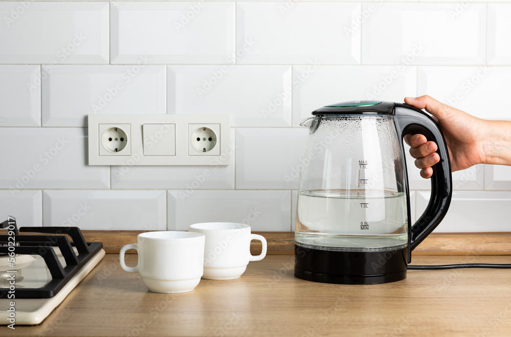 A transparent electric kettle with boiling water in a woman's hand and cups for tea on the table in the kitchen. The woman is making tea. A transparent electric kettle with boiling water in a woman's hand and cups for tea on the table in the kitchen. The woman is making tea.